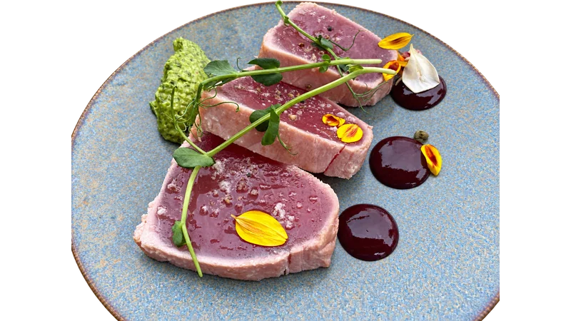 Seared tuna with pea shoots, yellow petals and sauce on a blue ceramic plate.