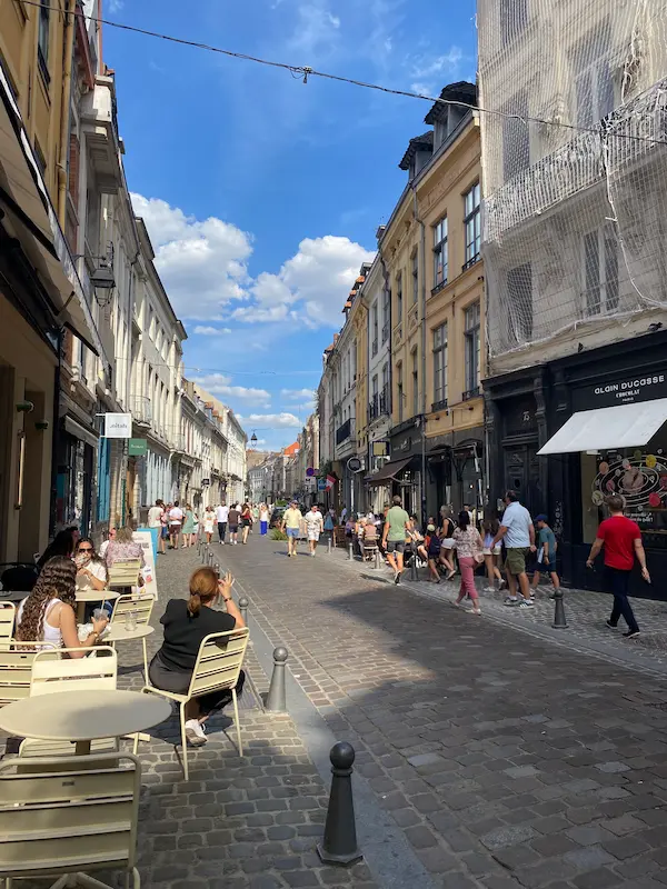 Pedestrians strolling along Rue Basse in Old Lille, a cobbled street lined with historic buildings, shops and café terraces.